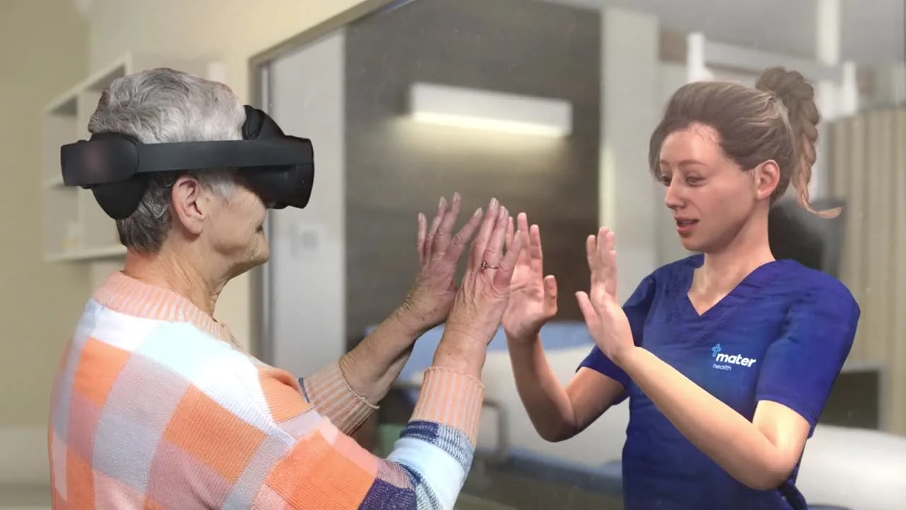 An older woman wearing a VR headset stands face to face with a nurse in Mater scrubs, both with their hands raised, in a simulated hospital room.
