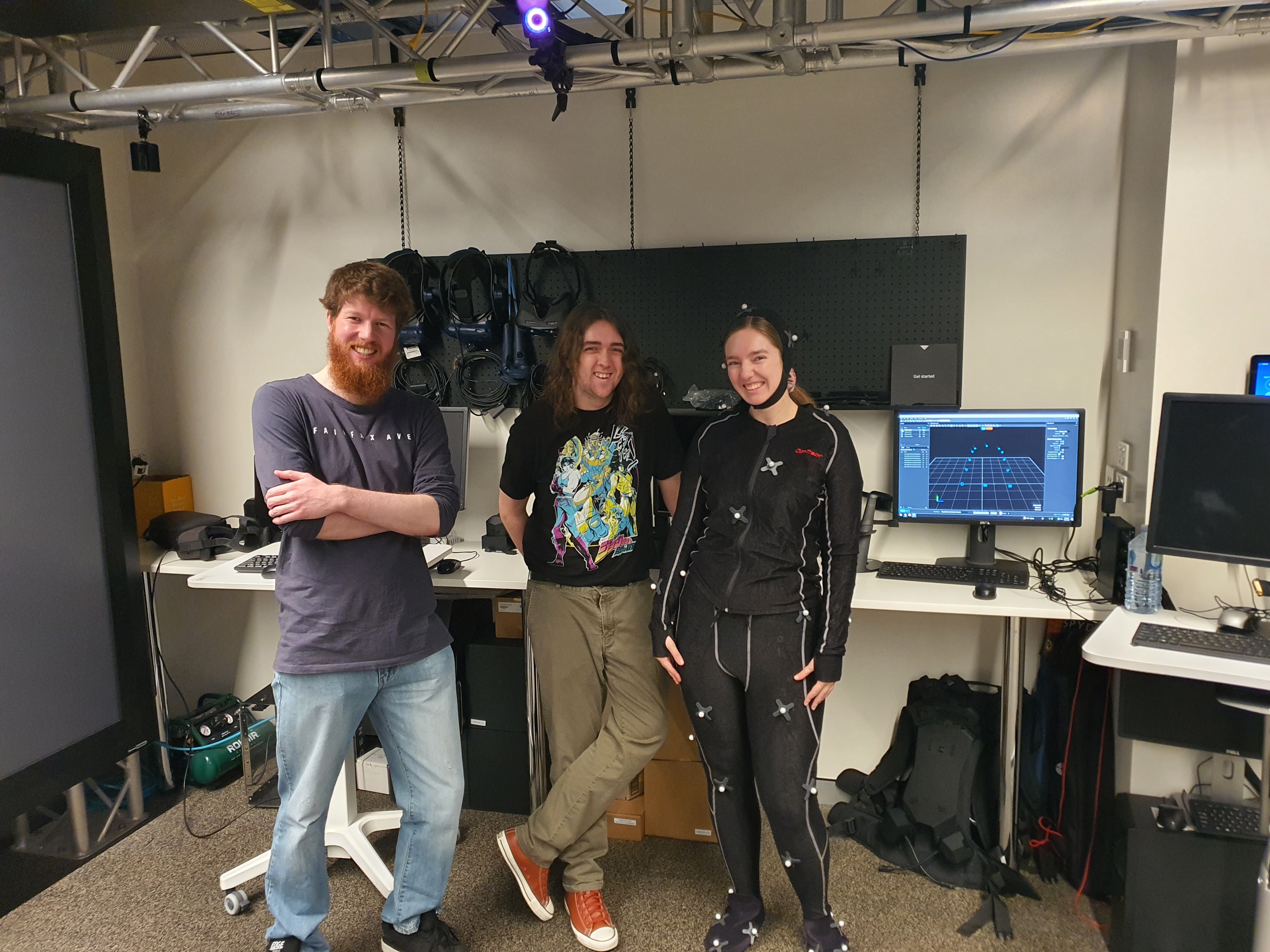 Three PTR crew members in a motion-capture lab, one wearing an OptiTrack mocap suit, with VR headsets and monitors behind them