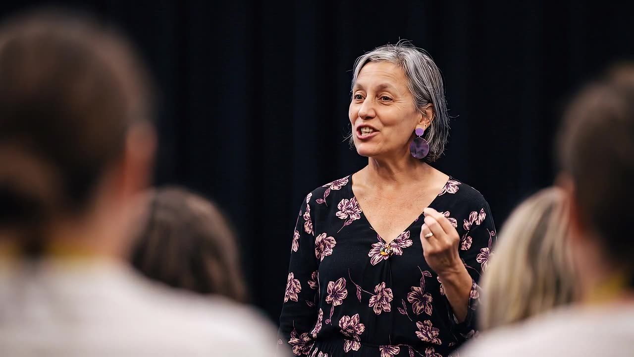 A PTR co-founder speaks to a seated room, gesturing mid-sentence, the audience soft-focused in the foreground as she explains the method behind the practice.