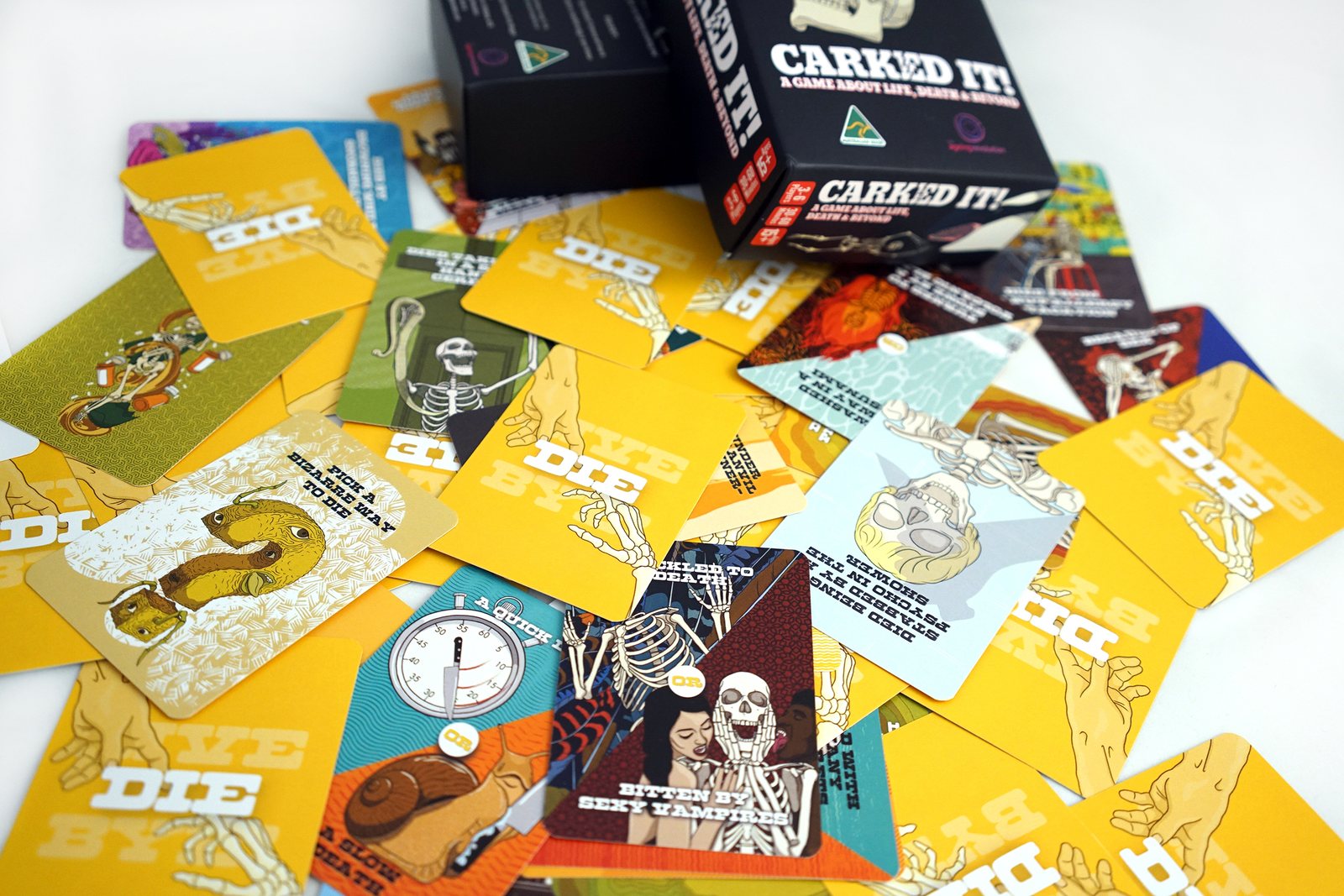 Yellow cards from the DIE deck fanned across a table beside the box, each illustrated with a different way of dying and titled in white capitals.