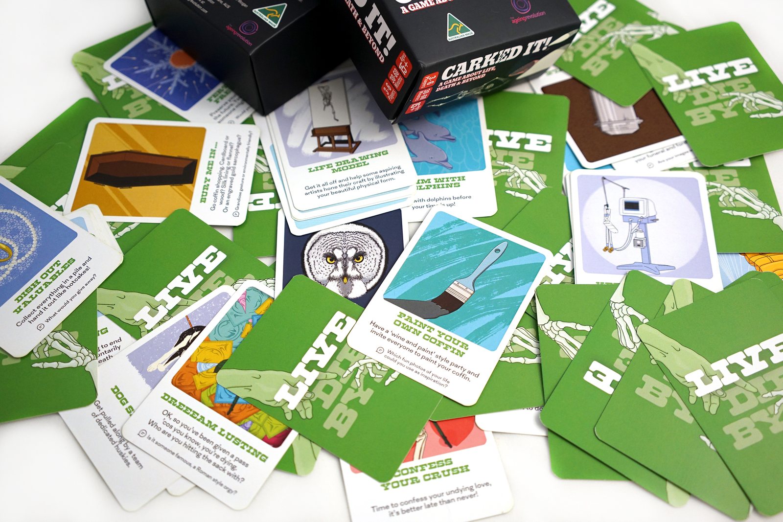 Green cards from the LIVE deck spread face up around the box, illustrated with a life drawing easel, a paintbrush on a coffin and a diver, each card carrying a small printed task.