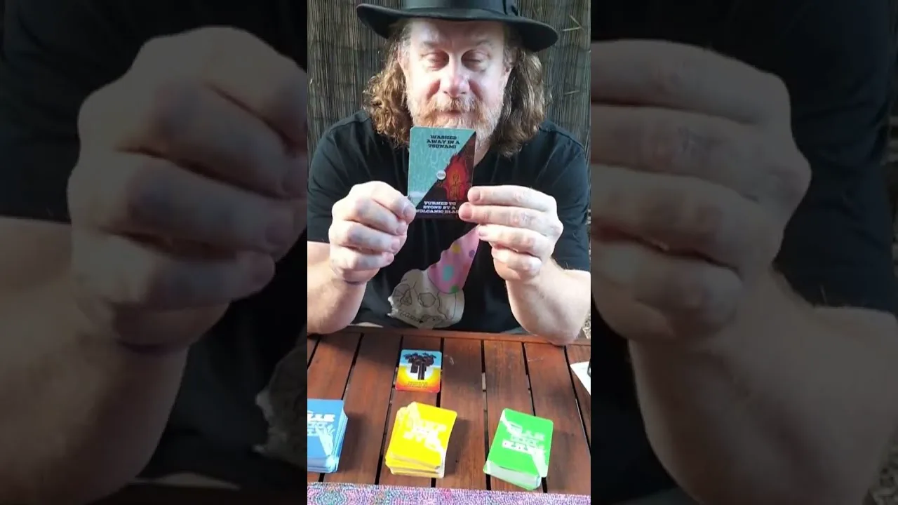 A man in a wide-brimmed hat holds a card up to camera above a table of Carked It! decks, the opening frame of the rules video.
