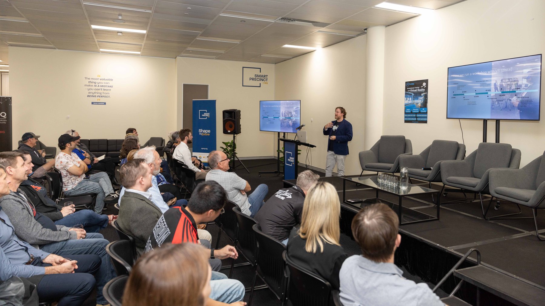 A seated audience watches a presenter explain a slide titled 'How we run a workshop with VR' at a PTR community event