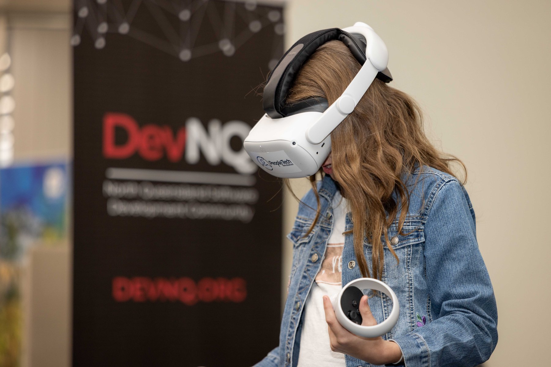 A young woman in a denim jacket wearing a PeopleTech Revolution branded VR headset, holding a controller, at a PTR community event