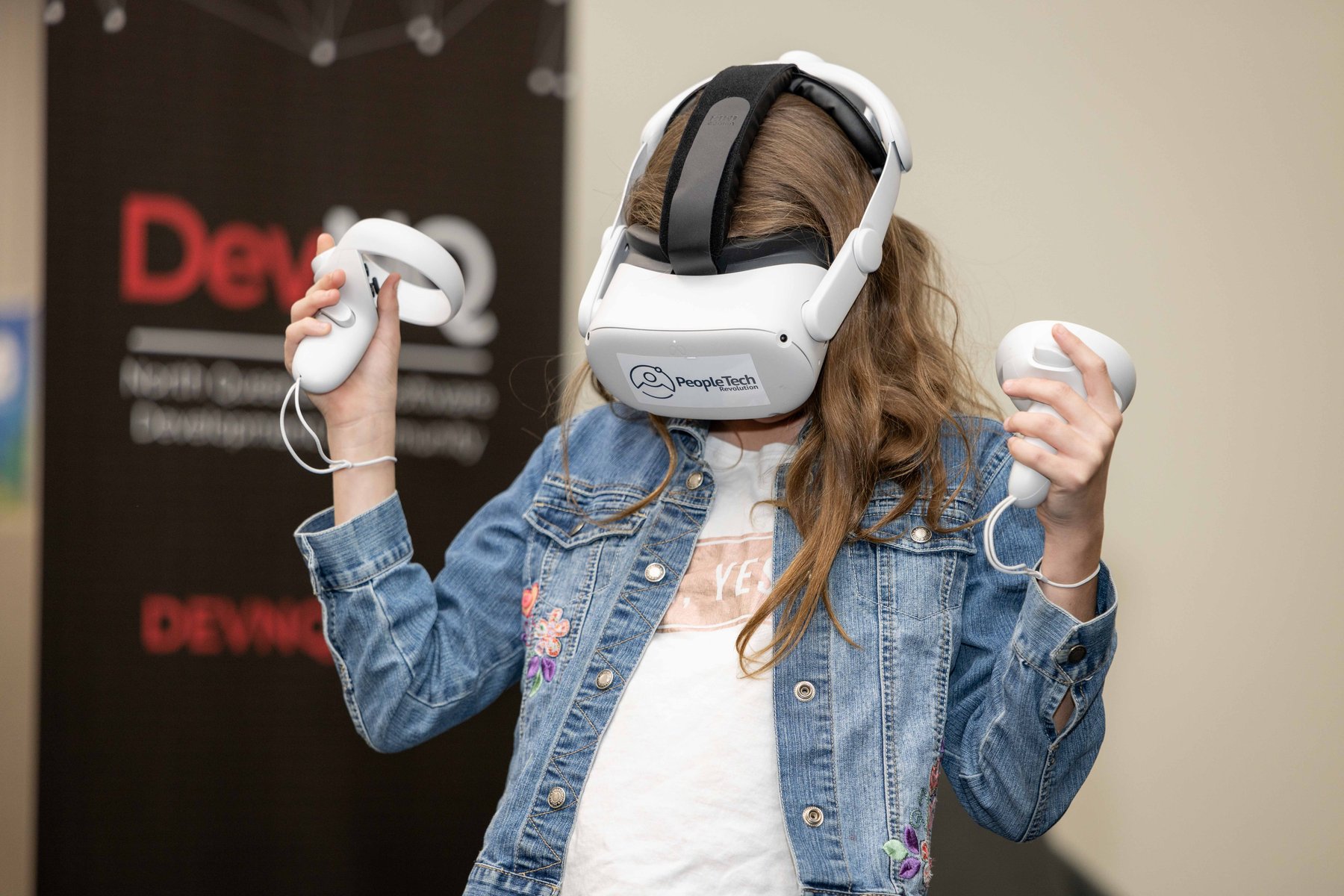 A young woman in an embroidered denim jacket wearing a PeopleTech Revolution branded headset raises both controllers at a PTR community event