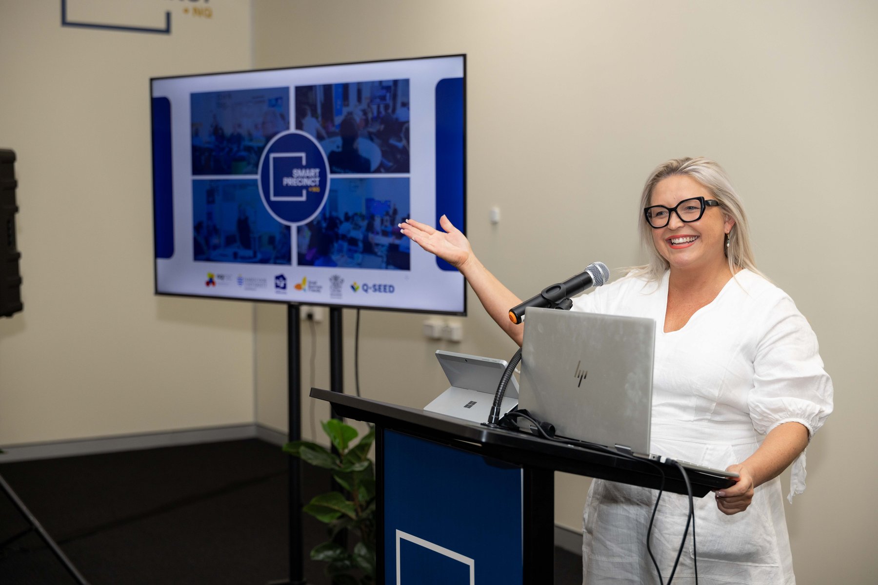 A smiling presenter in glasses gestures toward a slide screen while speaking at a lectern during a PTR community event