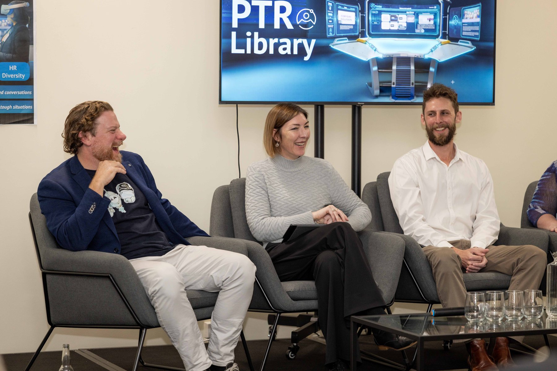Three panellists laughing together in armchairs beneath a PTR Library screen at a PTR community event