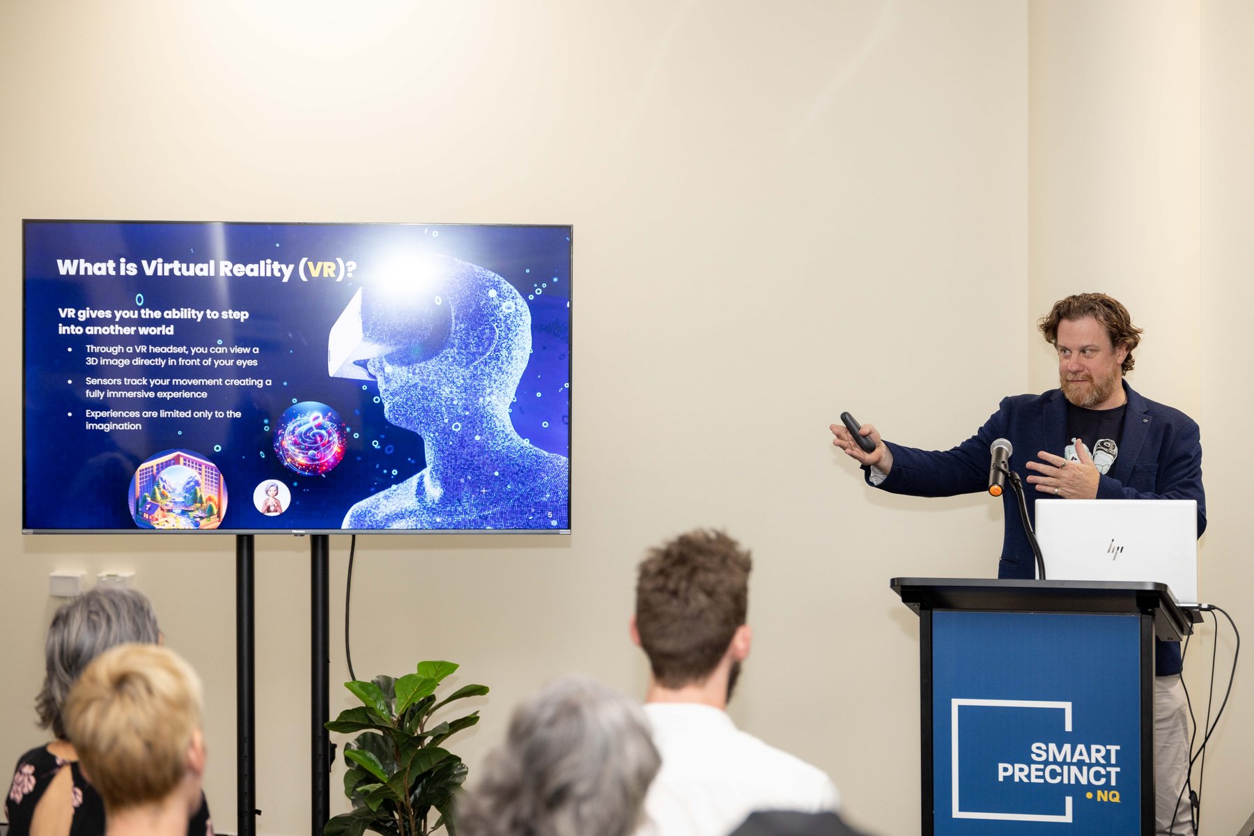 Simon Lowe presents beside a screen showing a 'What is Virtual Reality (VR)?' slide to a seated audience at a PTR community event