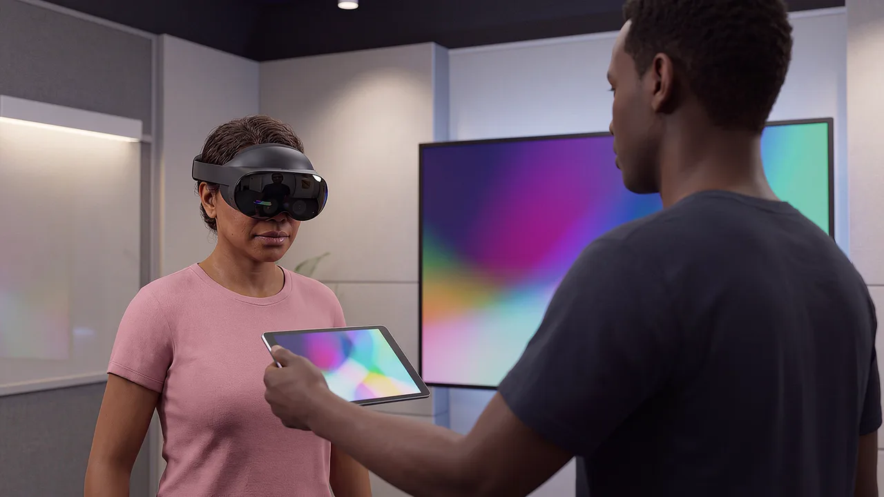 A man holds out a tablet showing a working scene to a woman in a headset, in a studio with a large screen behind them.
