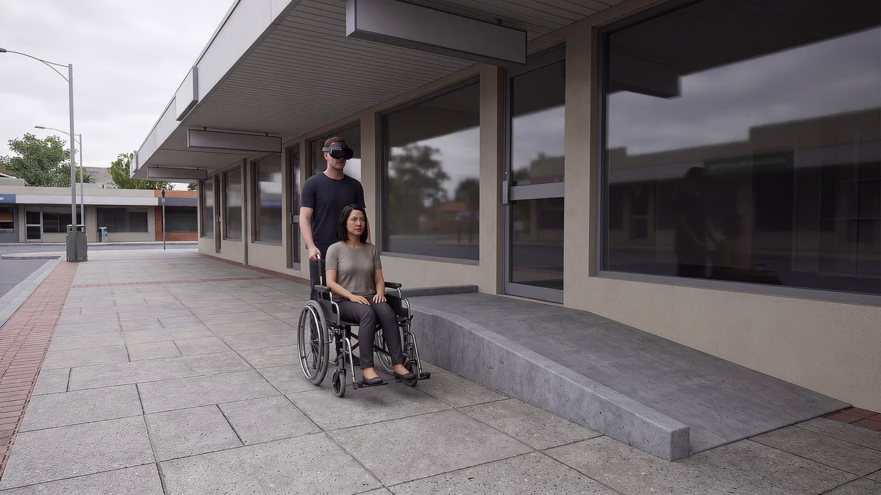 A simulated shopping strip with a ramp to a doorway. A woman waits in her wheelchair at the ramp while a support worker wearing the headset stands beside her.