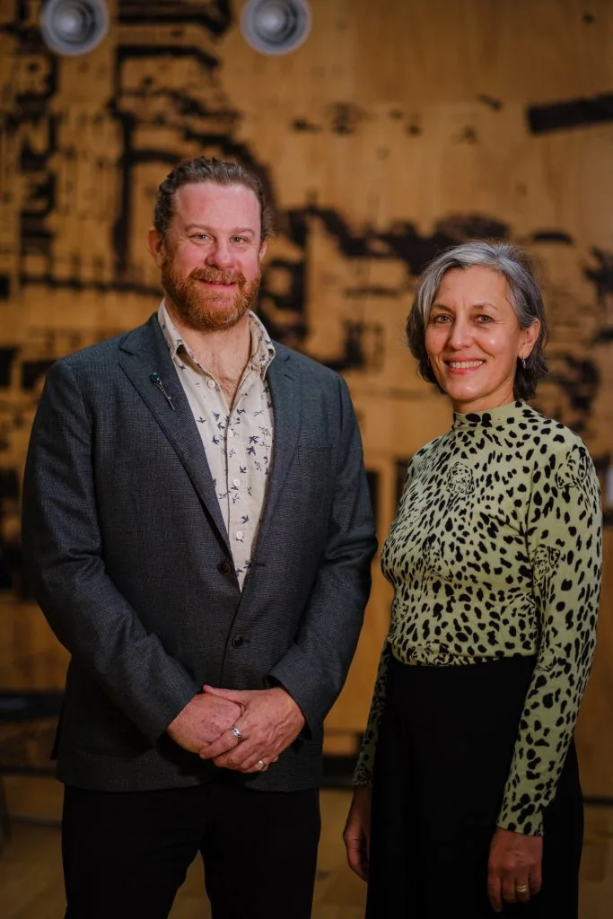 Simon Lowe in a grey blazer and Leonie Sanderson in a green patterned top, standing together in front of a dark wood-burnt wall.