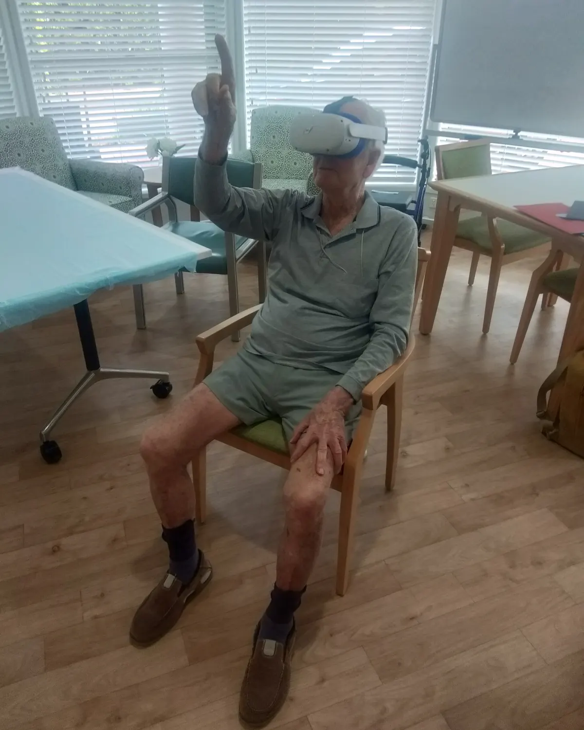 An older man wearing a VR headset raises one arm and points upward, seated in a wooden chair in a care home day room.