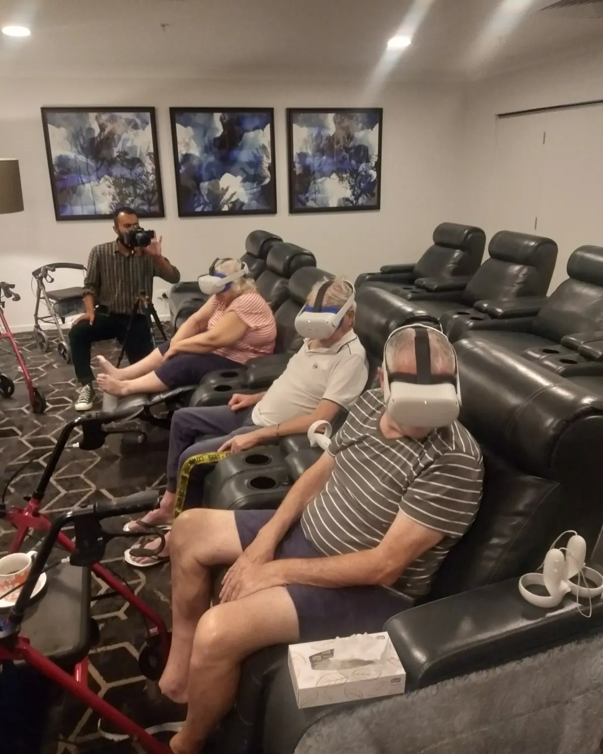Older residents in a row of black recliners, some wearing white VR headsets, while a man behind them films the session with a camera on a tripod.