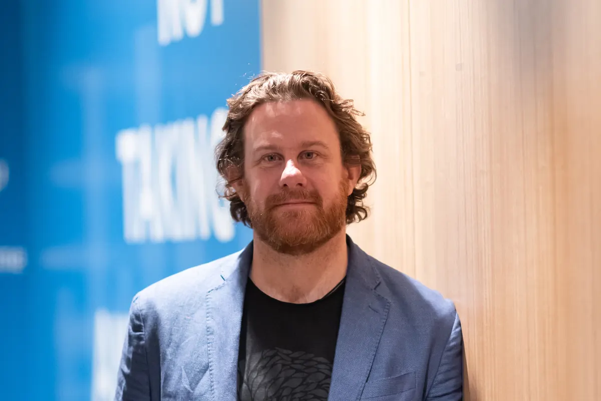 Simon Lowe in a blue blazer and dark t-shirt, smiling, against a timber wall.
