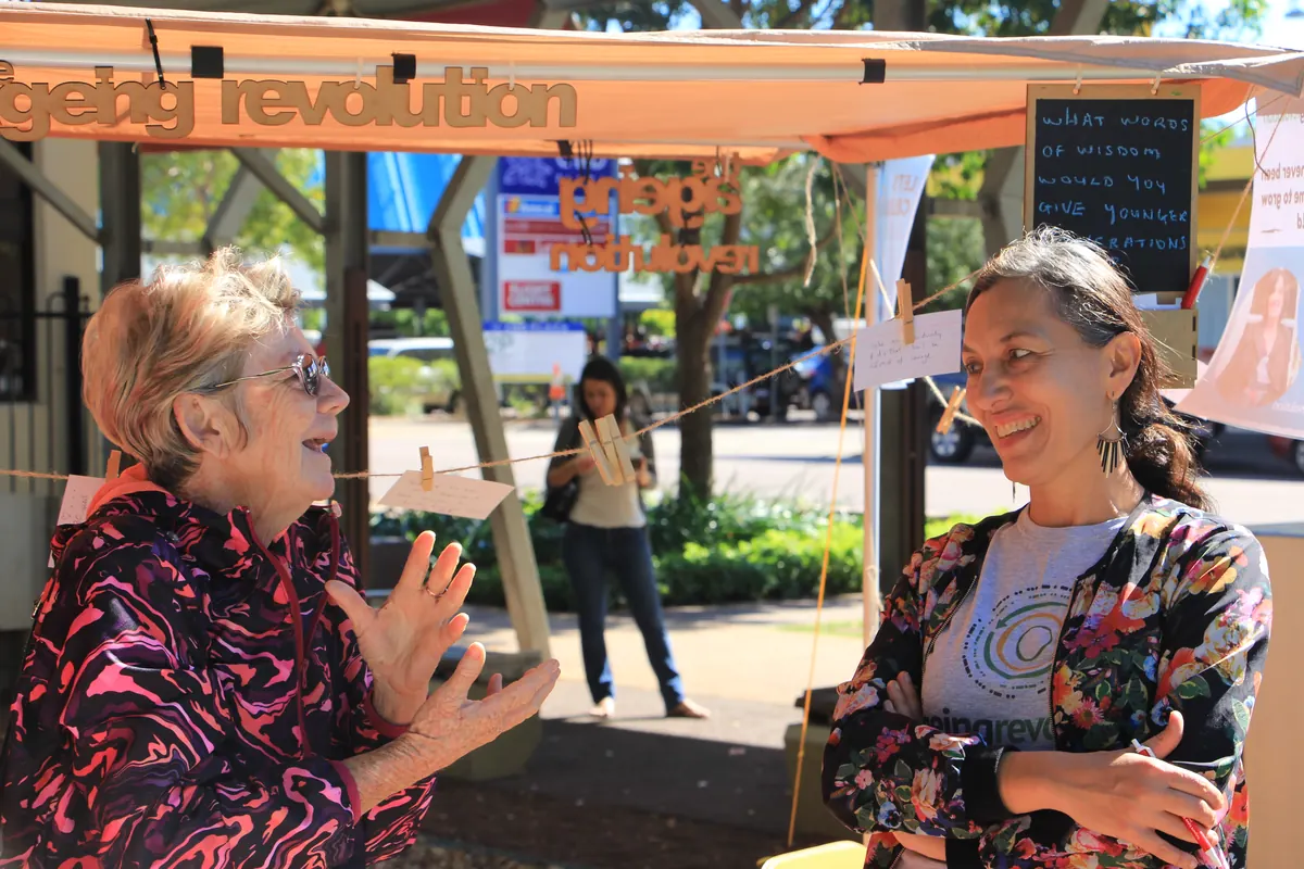 Leonie Sanderson, in an Ageing Revolution shirt, laughs with an older woman beside the stall, a chalkboard behind them asking for words of wisdom for younger generations.