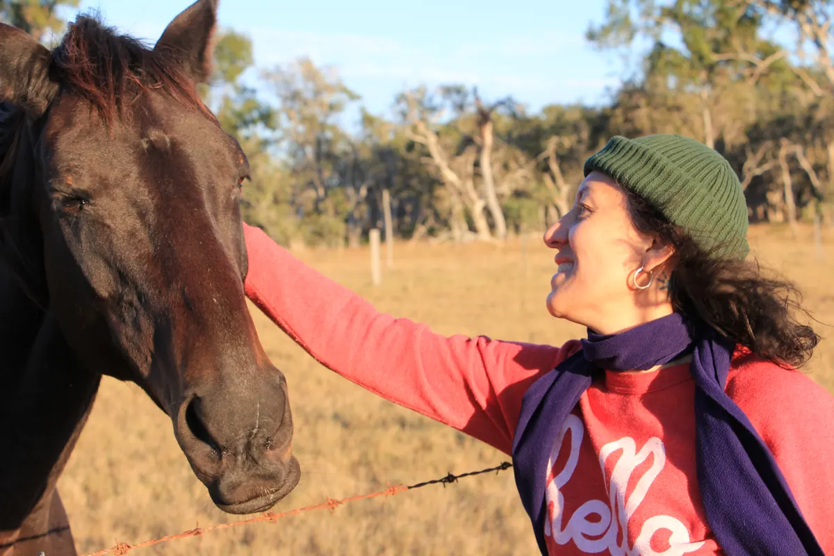 Leonie Sanderson in a beanie and scarf reaches over a fence to stroke a horse's face in a dry paddock.