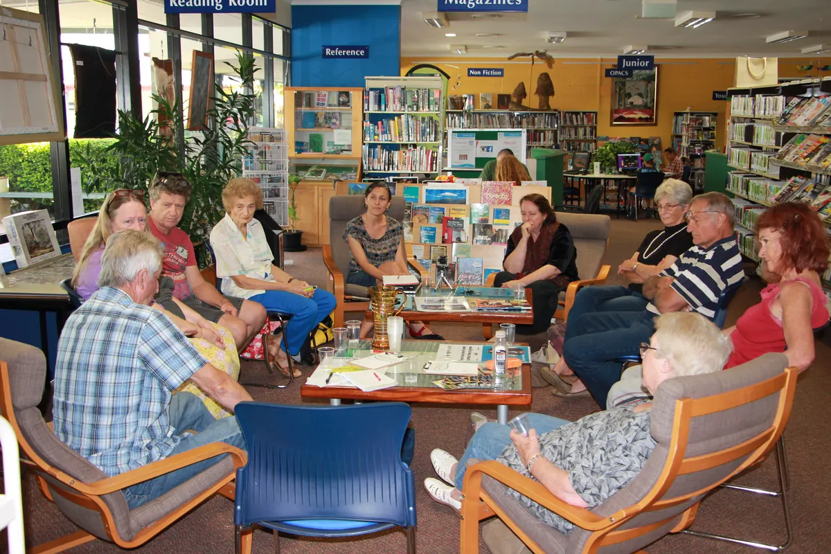Older people sit in a circle of armchairs in a library, listening as one of them speaks, with Leonie Sanderson seated among them.