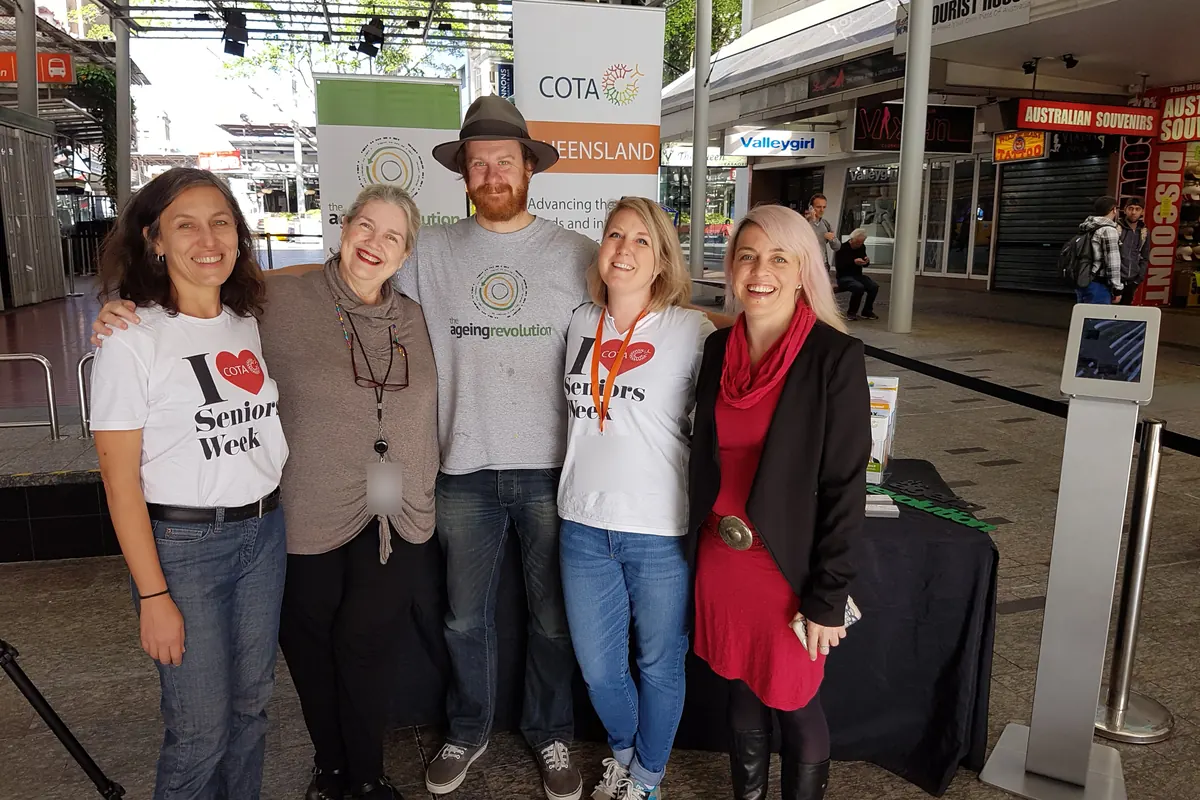 Leonie Sanderson and Simon Lowe stand with three others at a stall in a shopping mall, under COTA Queensland and Ageing Revolution banners; two of them wear I love Seniors Week shirts.
