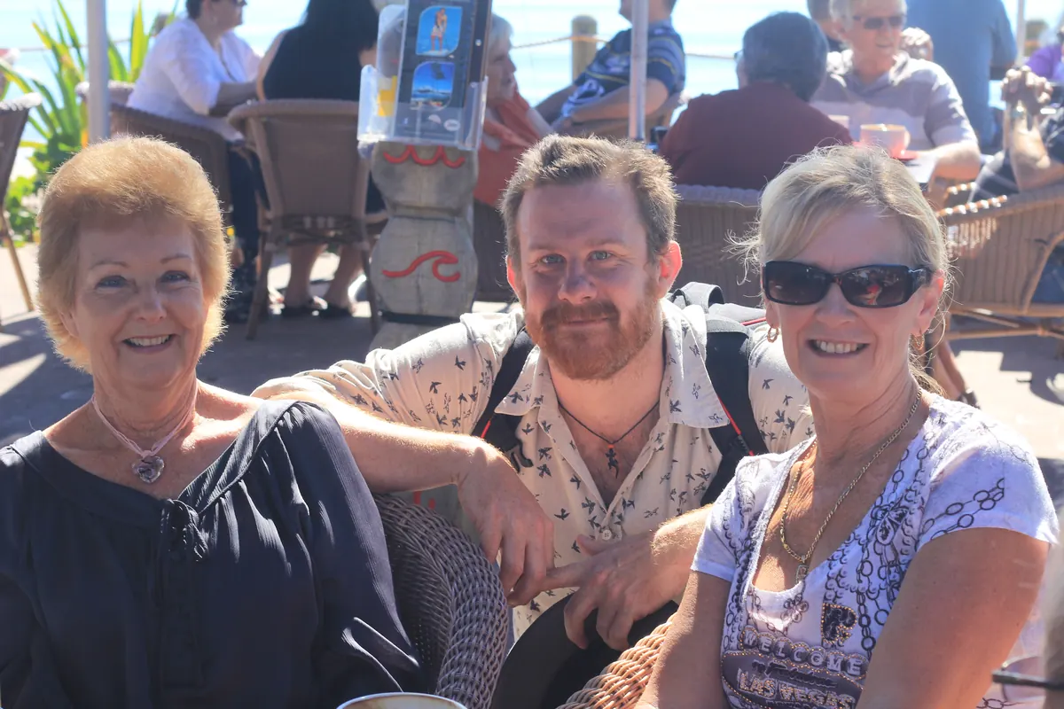 Simon Lowe smiles beside older women at an outdoor seaside cafe table.
